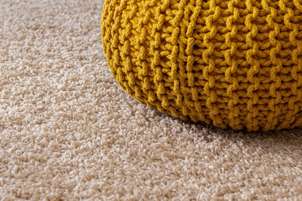 A picture of a light-colored, soft, and fluffy carpet with a mustard crochet cushion.